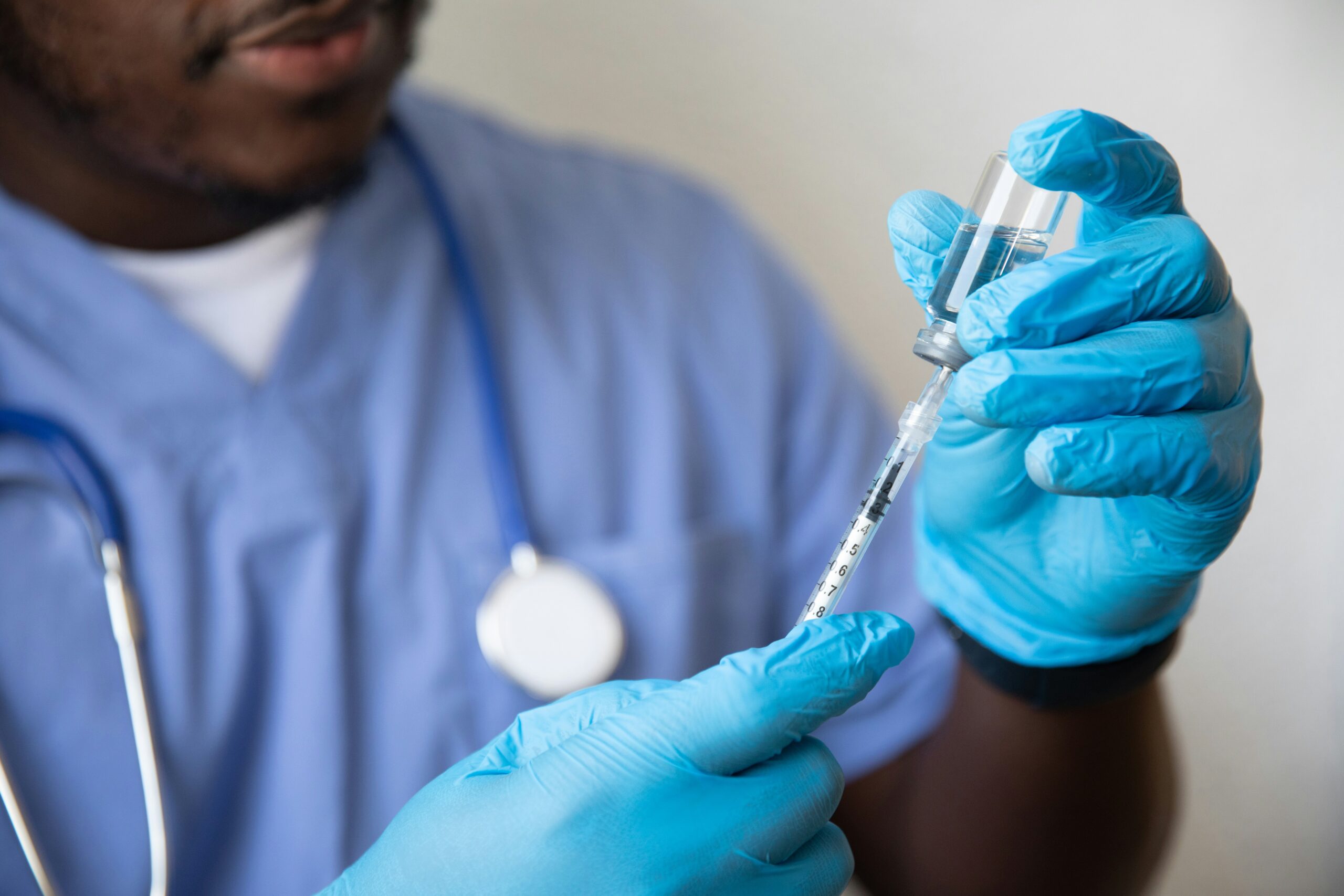 A man in blue scrubs and gloves holds a syringe to a glass vial
